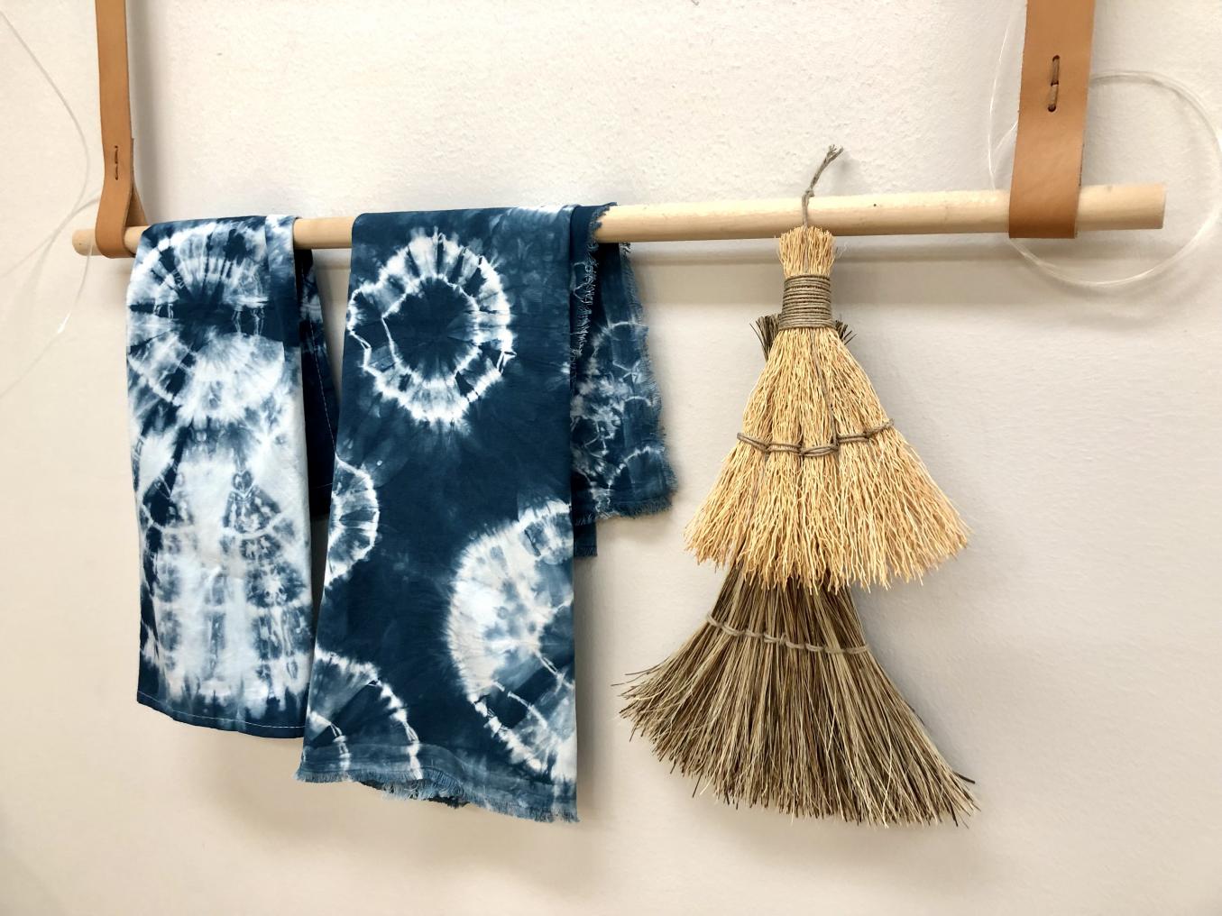 Kaksi shibori-värjättyä liinaa ja viskaa roikkuu vaakatasoon asetetusta puukepistä.